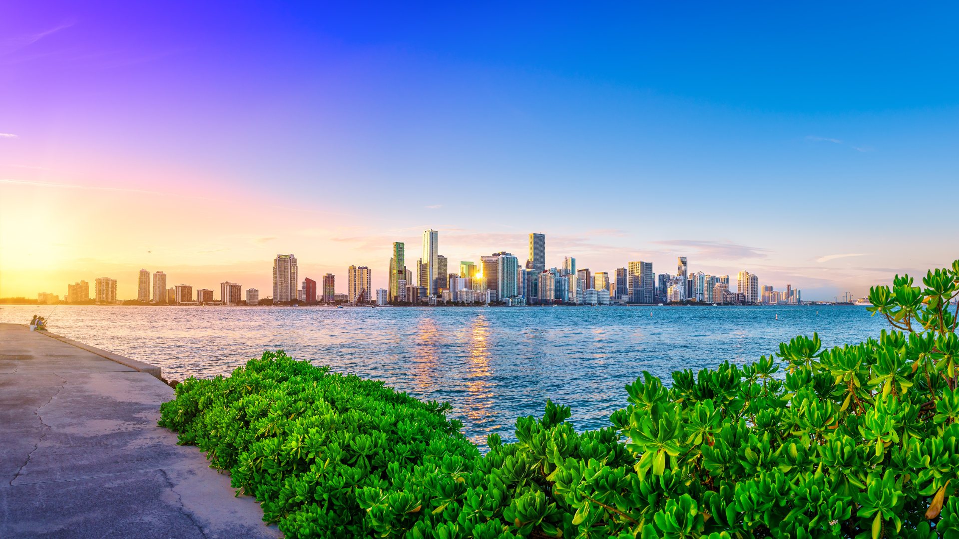 Miami skyline at sunrise across Biscayne Bay, seen from a waterfront promenade lined with coastal greenery